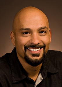 A man with tan skin and a dark beard wearing a button-down shirt smiles for a portrait.