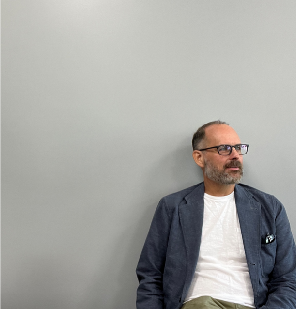 A man with short salt-and-pepper hear and a beard wearing glasses sits against a gray wall. 