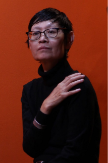 A person with short, dark hair and glasses wearing a long sleeve black sweater, their arms wrapped around themselves