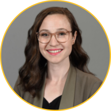 Woman with dark, wavy hair and pale skin wearing glasses and a brown suit jacket
