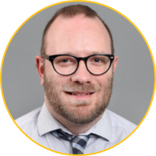 Man with short, light-brown hair and a beard wearing glasses and a tie